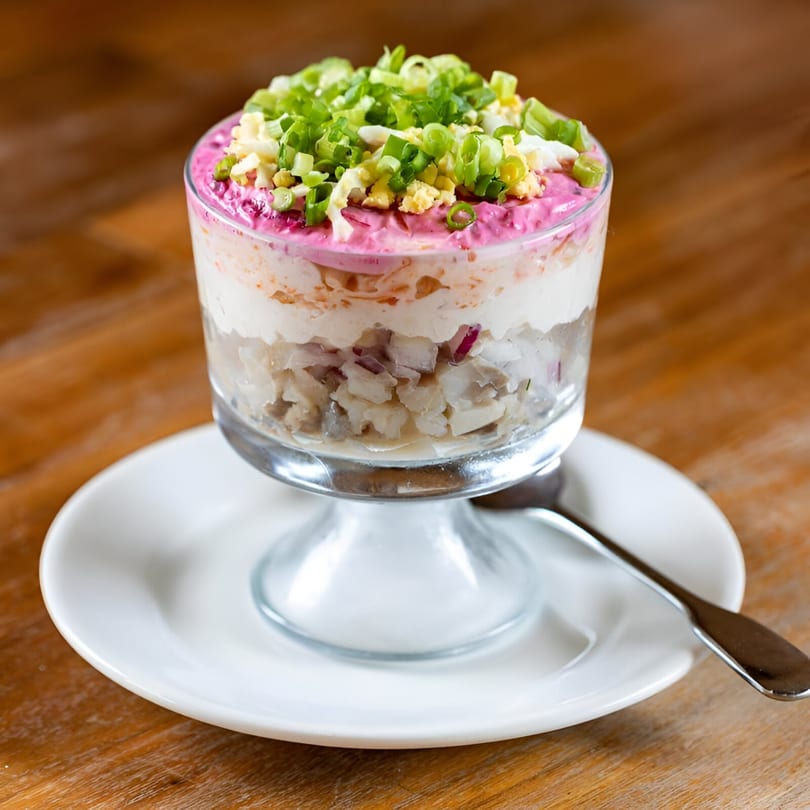 Layered European herring salad in a glass, topped with pink beet puree, chopped egg, and green onions.