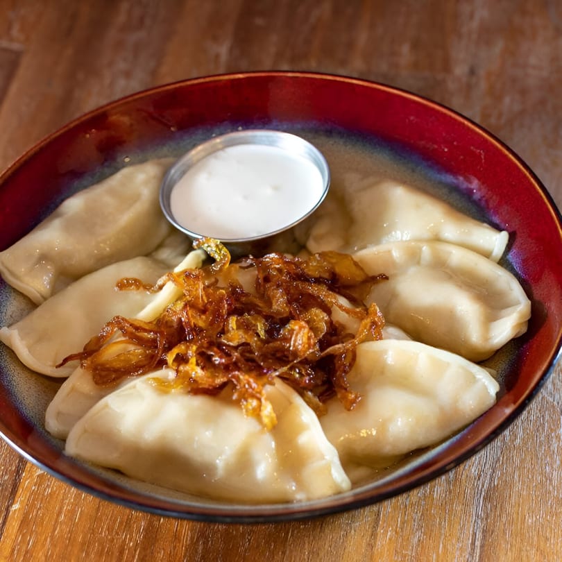 European dumplings topped with crispy fried onions and a side of sour cream in a red bowl.