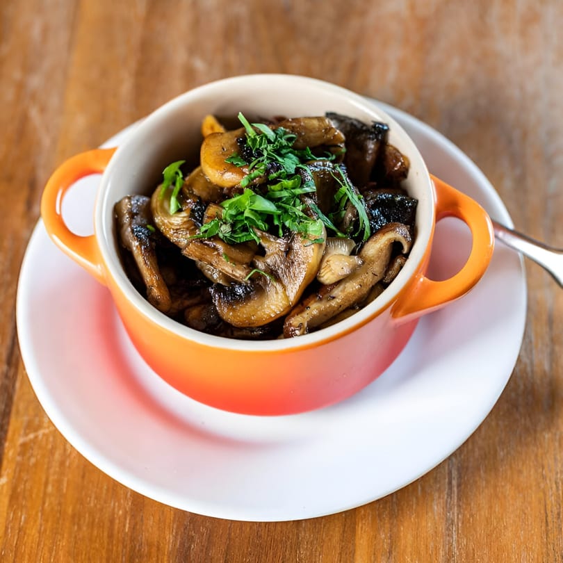 Sautéed mushrooms garnished with fresh herbs in an orange ceramic bowl on a white plate.