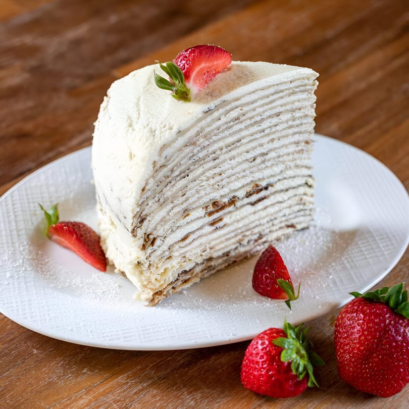 A slice of European crepe cake with many layers, topped with strawberries and powdered sugar.