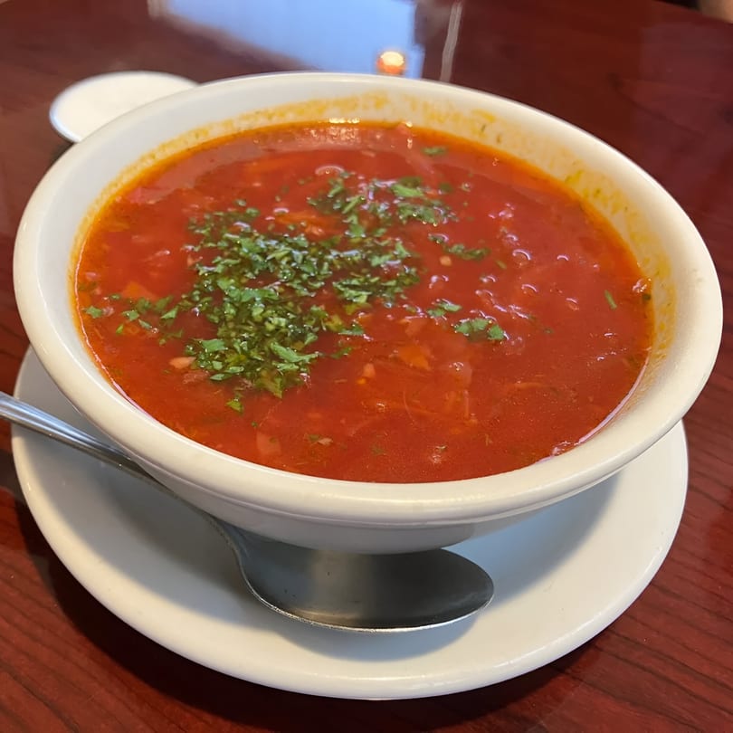 A white bowl of red borscht soup, garnished with fresh green herbs, on a wooden table.