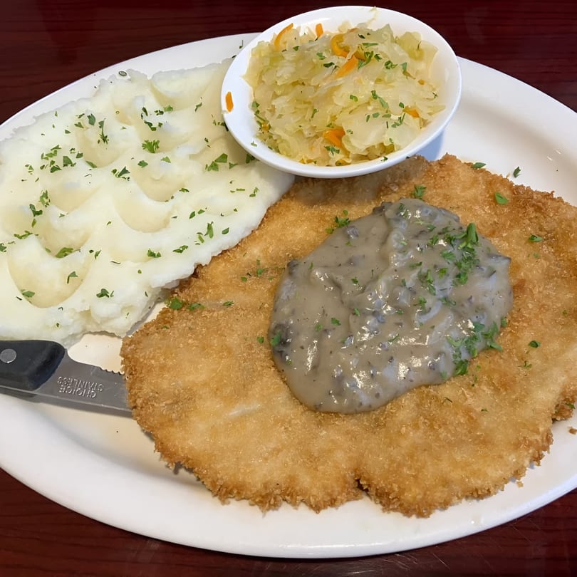 Golden-fried schnitzel with mushroom gravy, mashed potatoes, and sauerkraut on a white plate.